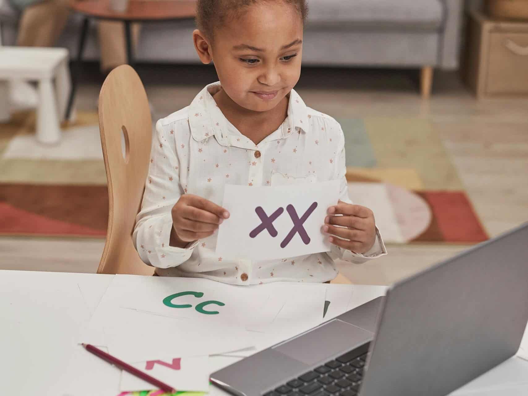 Child practicing alphabet through online learning with laptop at home. Mother watches in background.